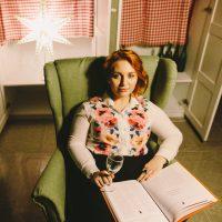 Lisa en una silla verde y con una copa de vino.